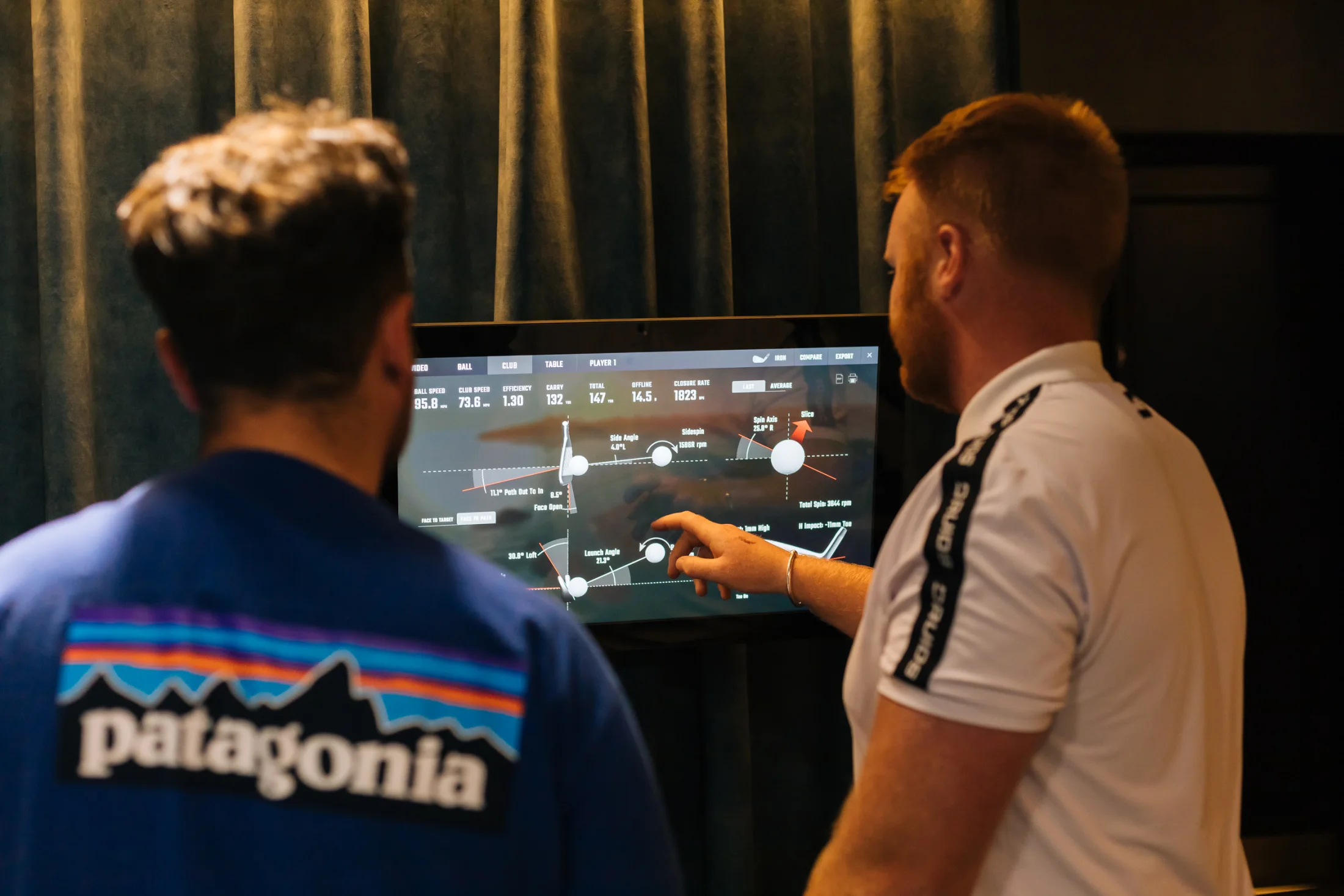 A Pitch coach explains shot and club data on a simulator screen to a golfer.