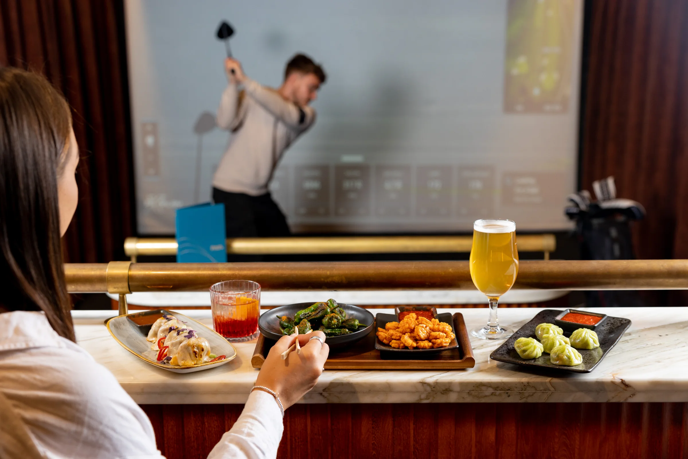 Food and drinks arranged at a Pitch simulator bay while a golfer plays in the background.