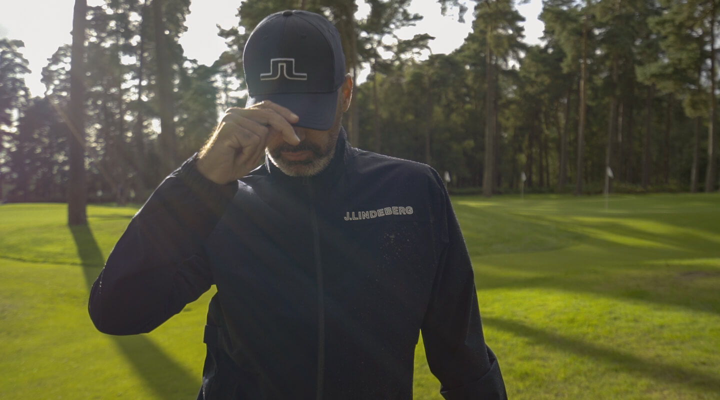 Golfer adjusting his cap while wearing a dark J.Lindeberg zip jacket in low sun at Woburn.